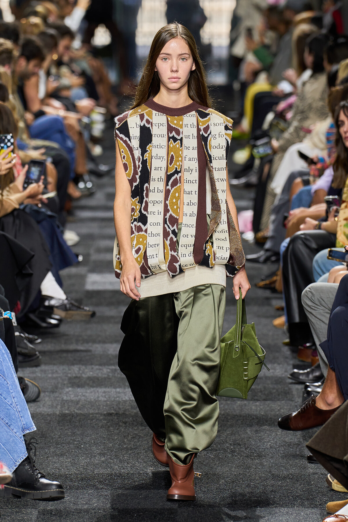 Model walks the runway in a bold, sleeveless patterned top with text, paired with olive-green satin pants, brown boots, and a green handbag.