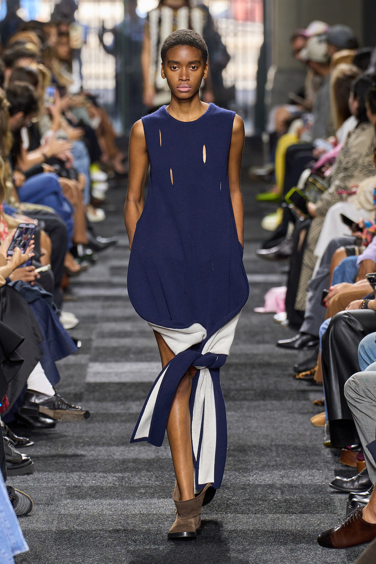 Female model walks a runway in a navy sleeveless dress with small cutouts and a navy-gray fabric wrap tied around her legs.