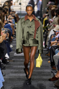 Model walking a fashion runway in an olive-green oversized coat with a brown leather collar, holding a yellow handbag.