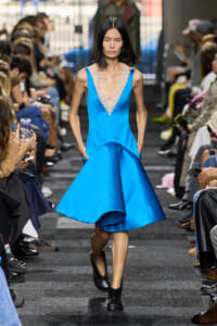Model walks the runway in a vivid blue satin dress with a deep lace V-neck and pockets, wearing black ankle boots.