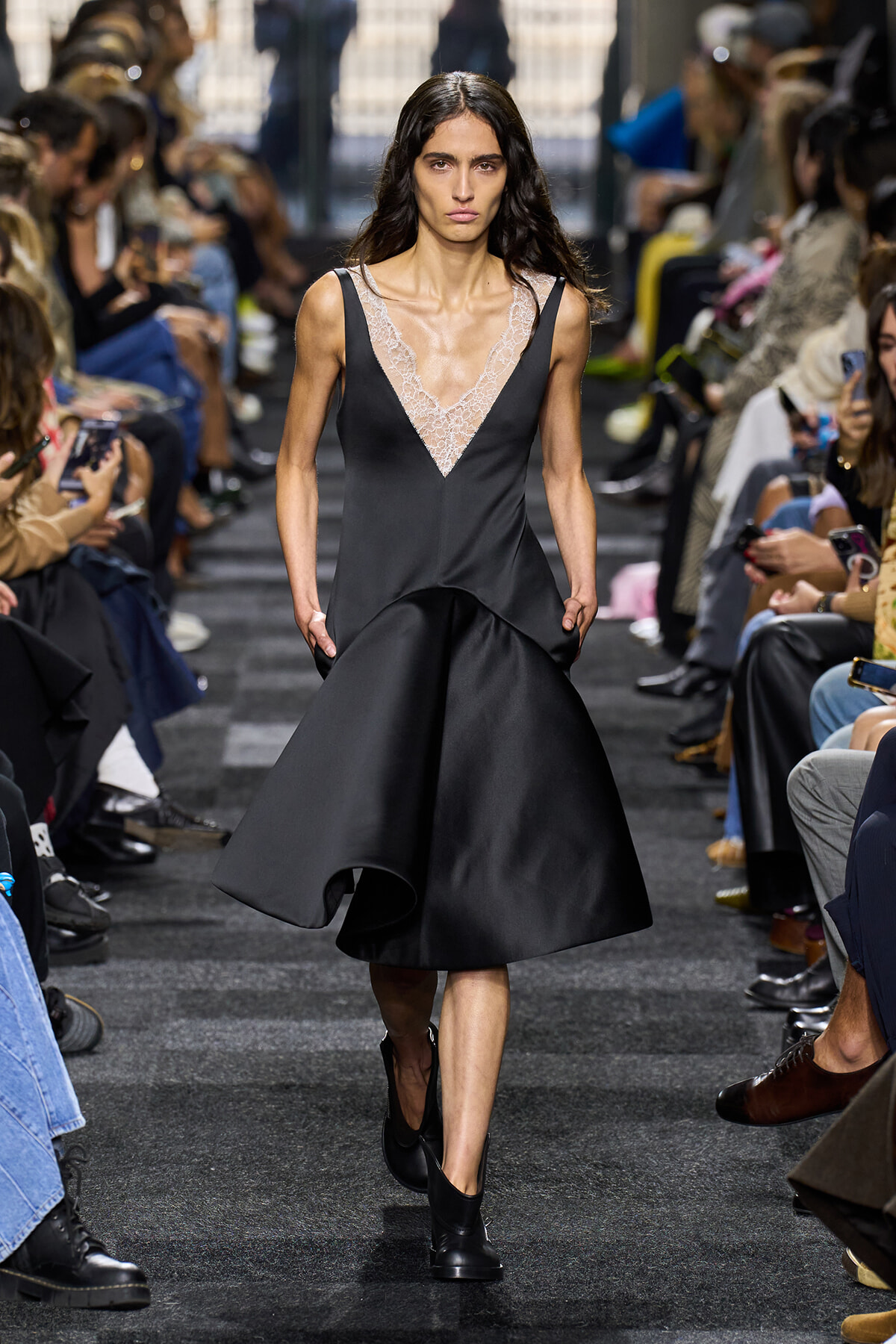 Model walks the runway in a black satin dress with a lace V-neck, hands in pockets, audience on both sides.