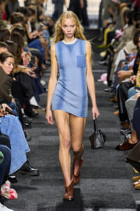 Model walks a fashion runway in a light blue sleeveless mini dress with a chest pocket, brown ankle boots, and a black handbag.