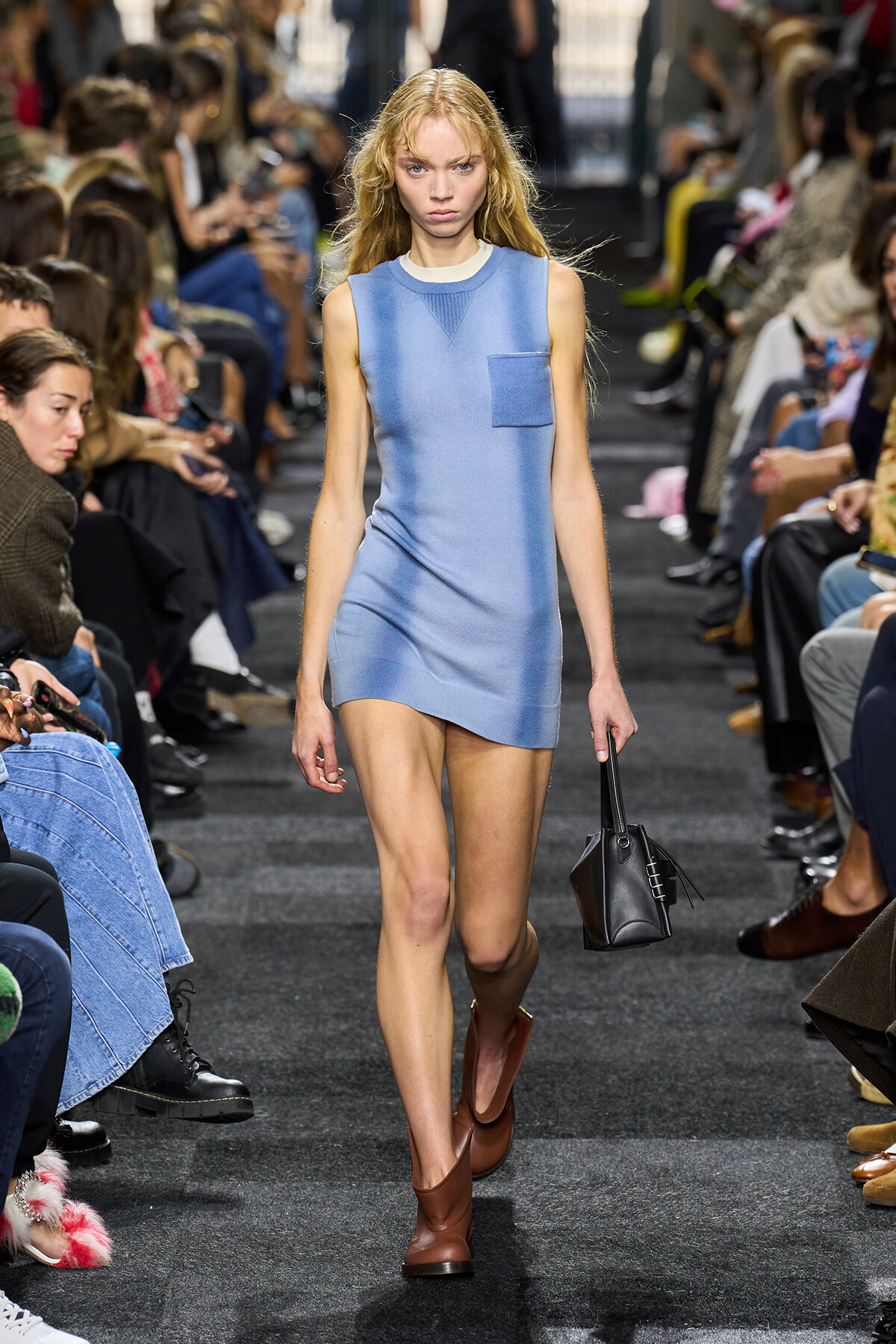 Model walks a fashion runway in a light blue sleeveless mini dress with a chest pocket, brown ankle boots, and a black handbag.