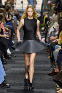 Model walks a fashion runway wearing a sleek black sleeveless dress with a wide, sculpted flare skirt. Audience sits on both sides in blurred focus behind her.