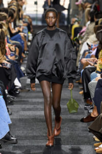 Model walks a fashion runway in a black oversized bomber, brown ankle boots, and a small green handbag.