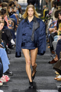 Model walks a fashion runway wearing a oversized navy jacket with black leather trim and matching shorts, black ankle boots, and a focused expression.