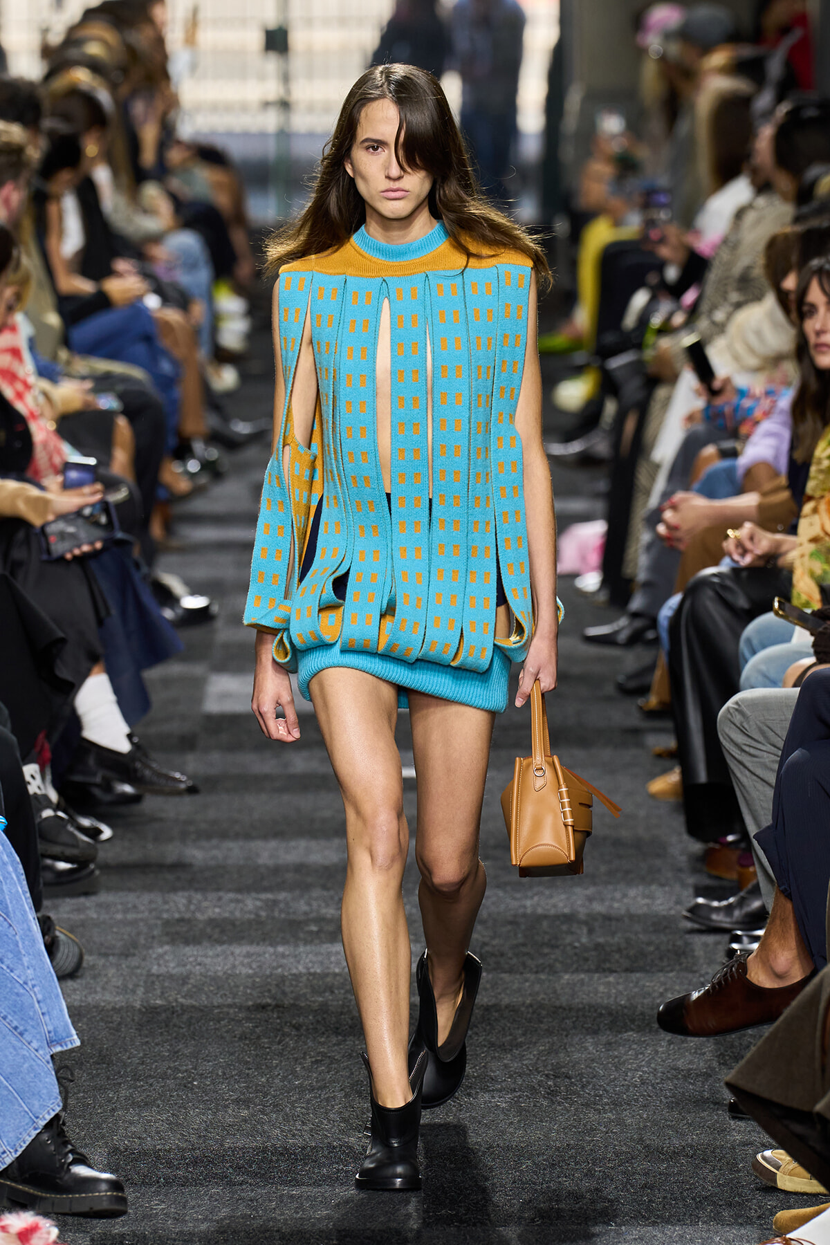 Model walks a fashion show runway in a turquoise knit dress with orange square patterns and a tan handbag.