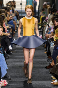 Model walks the runway in a mustard sleeveless top and a sculpted navy blue flared skirt, with brown ankle boots and seated audience along the catwalk.