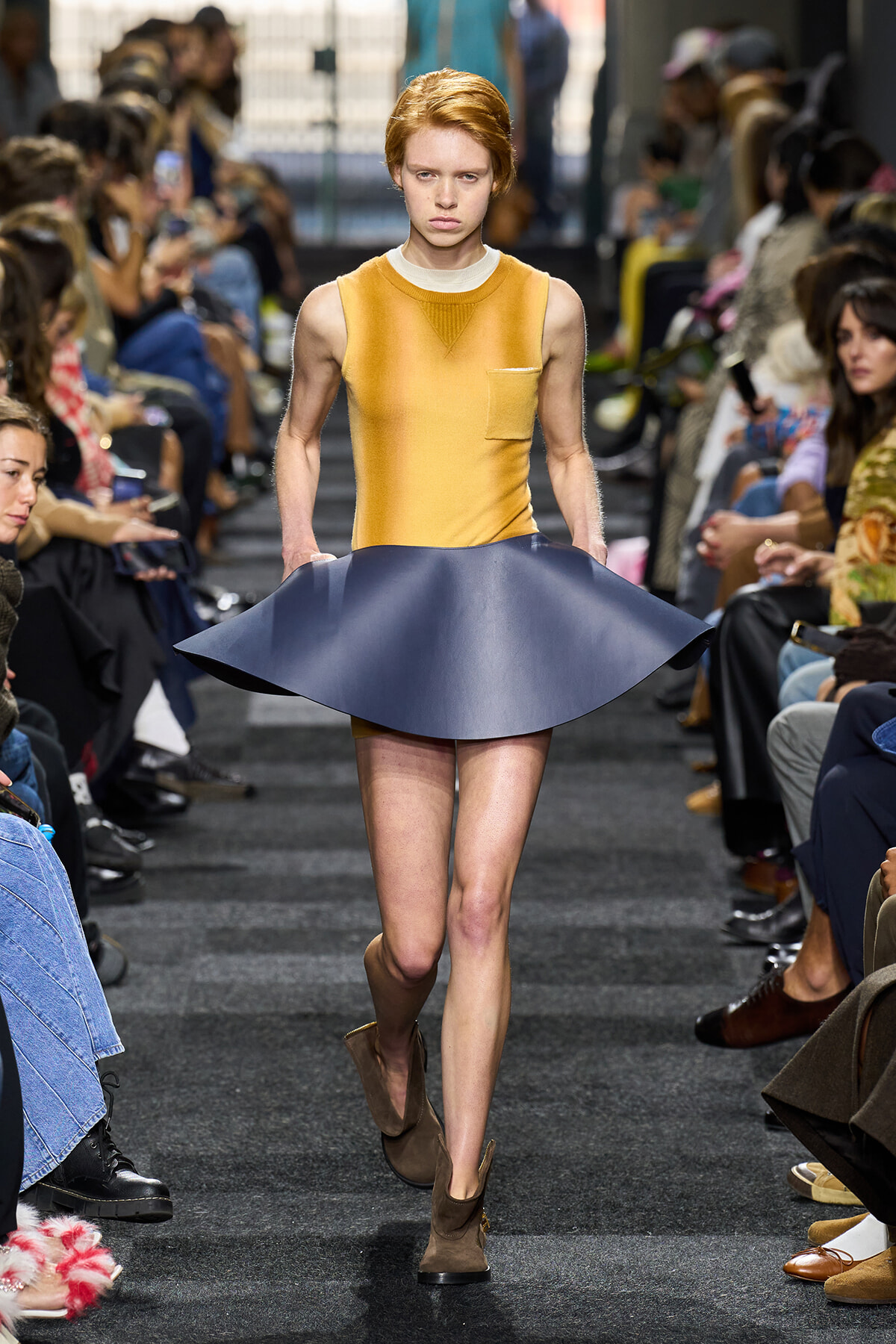Model walks the runway in a mustard sleeveless top and a sculpted navy blue flared skirt, with brown ankle boots and seated audience along the catwalk.