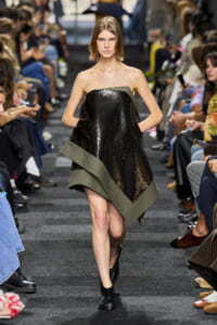 Model walks the runway in a strapless, asymmetrical black sequin dress with olive-green trim, surrounded by seated fashion show attendees.