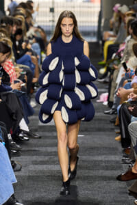 Model walks a runway in a navy knit dress made of stacked bubble-like segments with white interiors.