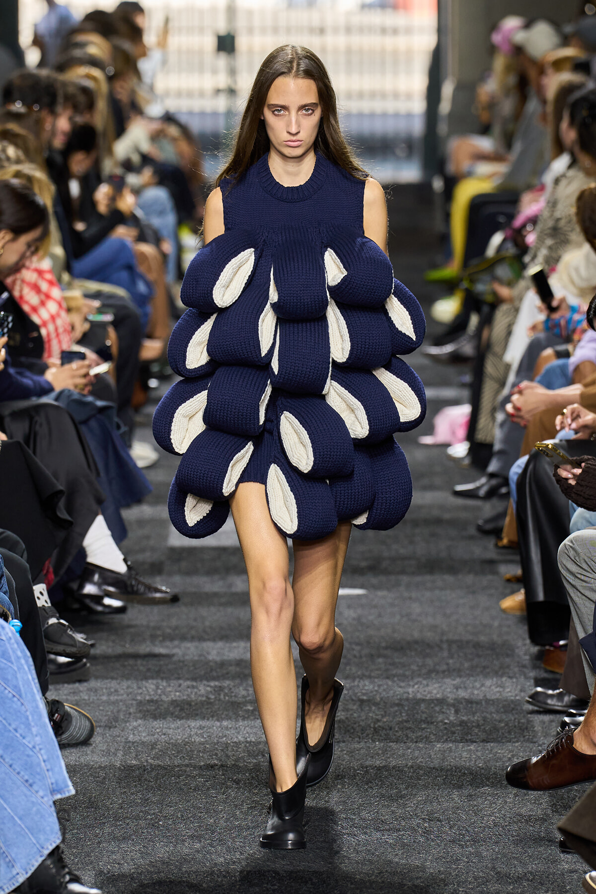 Model walks a runway in a navy knit dress made of stacked bubble-like segments with white interiors.
