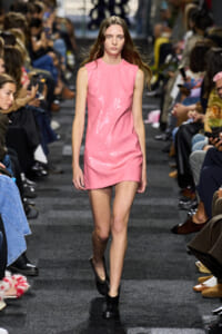 Model walks the runway in a pink sequined sleeveless mini dress, paired with black ankle boots, audience blurred in the background