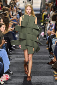 Model walks a runway wearing a sculptural olive-green knit dress with wide, overlapping bands wrapped around the torso and shoulders.