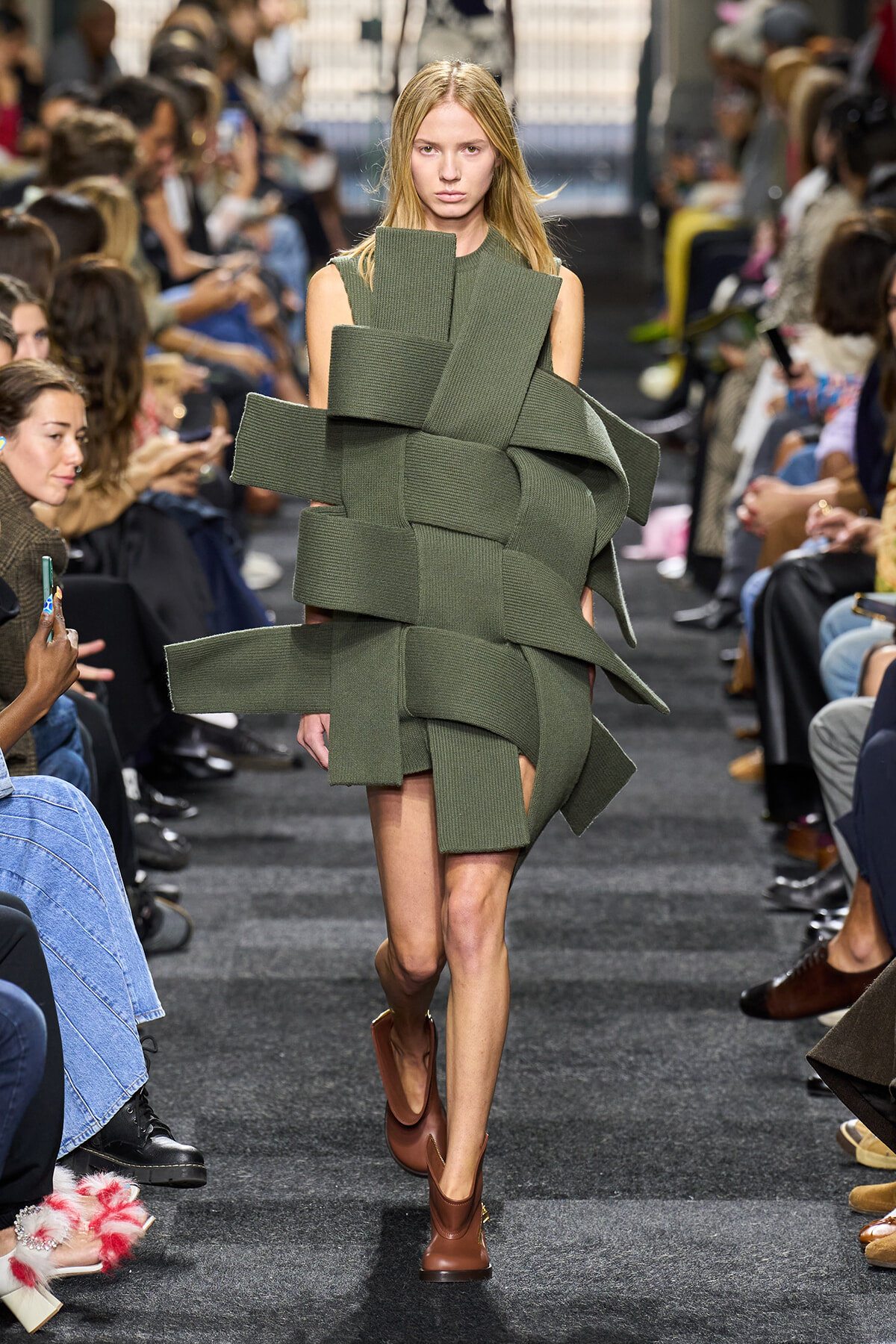 Model walks a runway wearing a sculptural olive-green knit dress with wide, overlapping bands wrapped around the torso and shoulders.