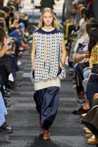 Model walks the runway in an oversized cream-and-navy knit with vertical cutouts, paired with navy satin pants and brown ankle boots.