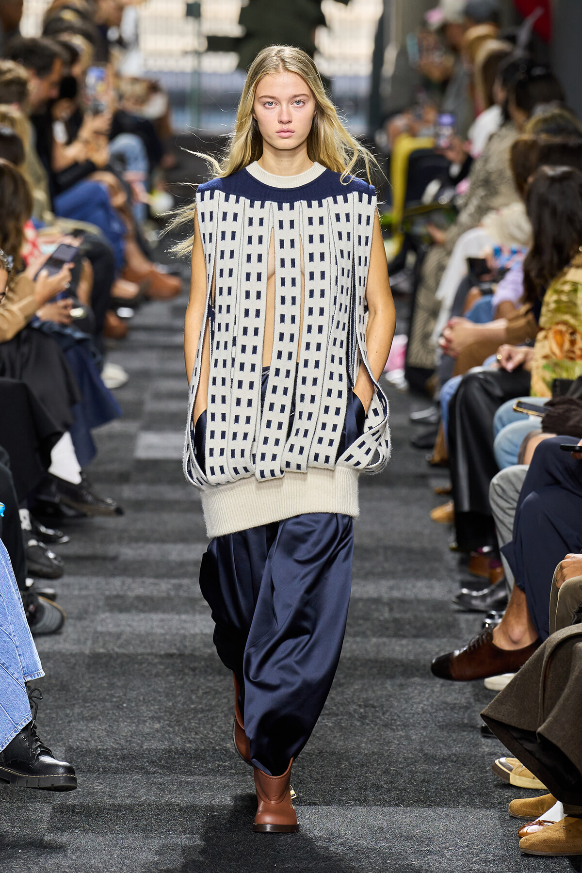 Model walks the runway in an oversized cream-and-navy knit with vertical cutouts, paired with navy satin pants and brown ankle boots.
