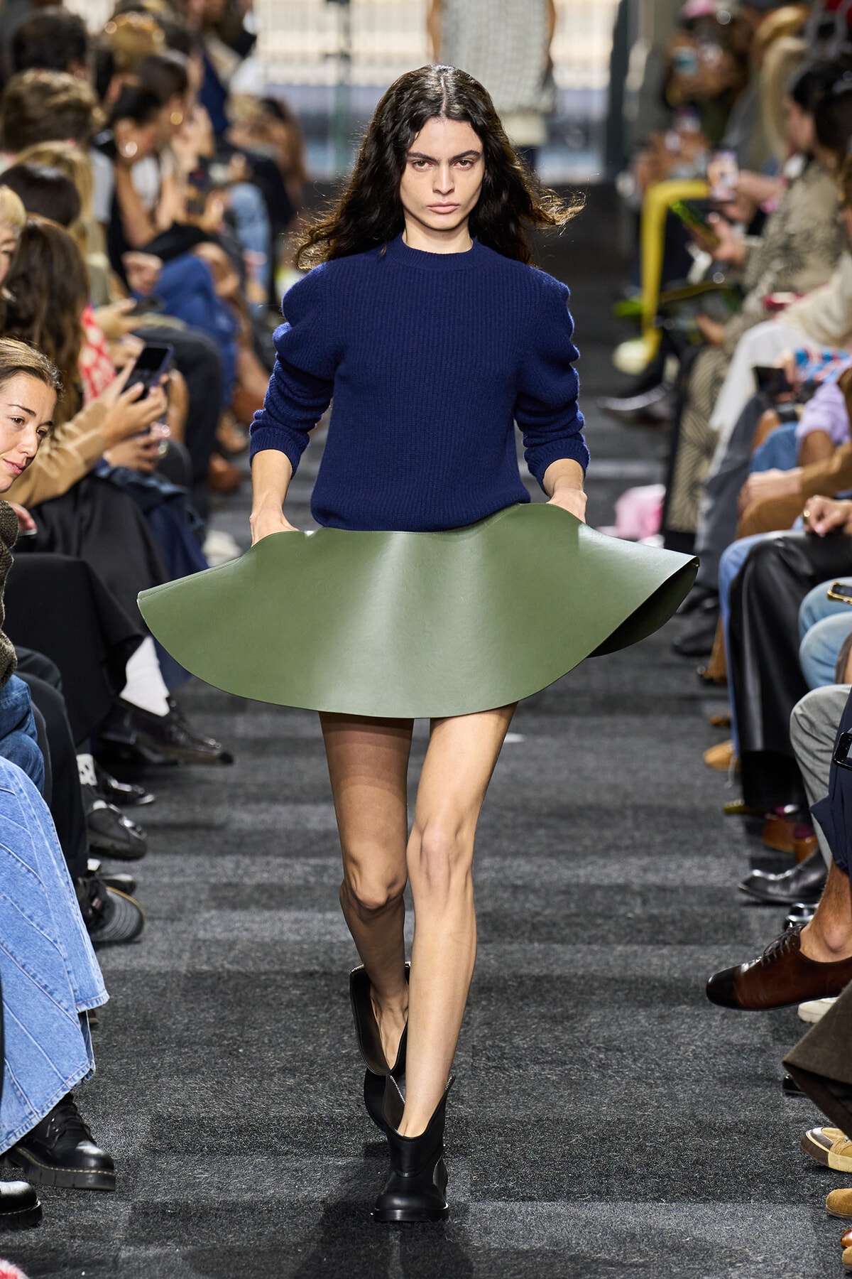 Model walks the runway wearing a royal blue knit sweater and a sculptural, curved green skirt that flips upward like a leaf under bright lights.