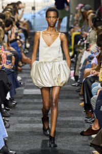 Model walks the runway in a cream satin V-neck top and a voluminous bubble skirt, paired with black ankle boots, as onlookers sit along the sides.