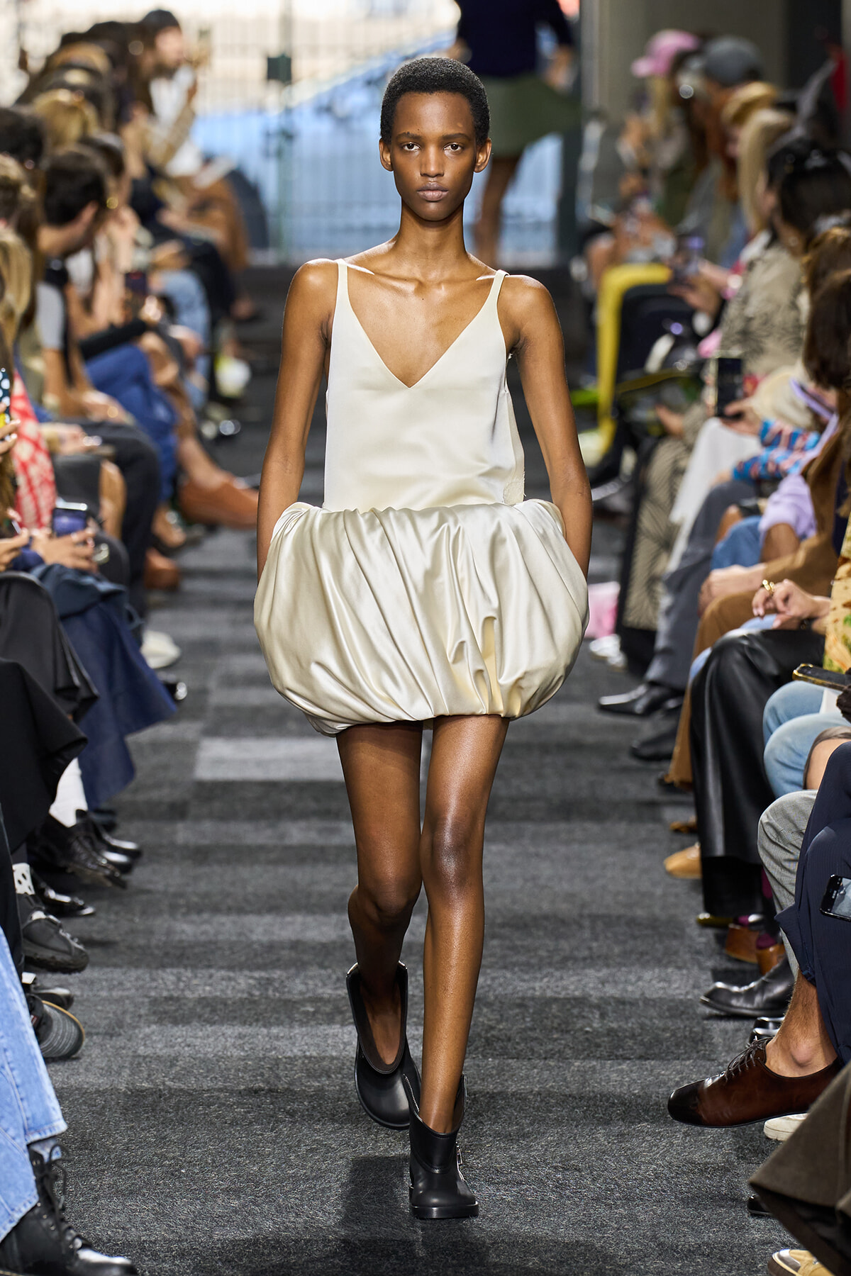Model walks the runway in a cream satin V-neck top and a voluminous bubble skirt, paired with black ankle boots, as onlookers sit along the sides.