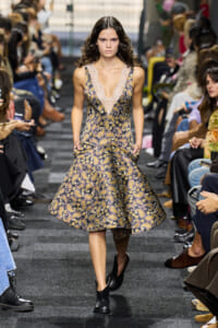 Model on a fashion runway wearing a sleeveless paisley dress with a deep lace V-neck and pockets, black ankle boots.