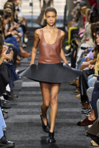 Model walks a fashion runway wearing a brown leather sleeveless top and a flared black skirt, audience on both sides.