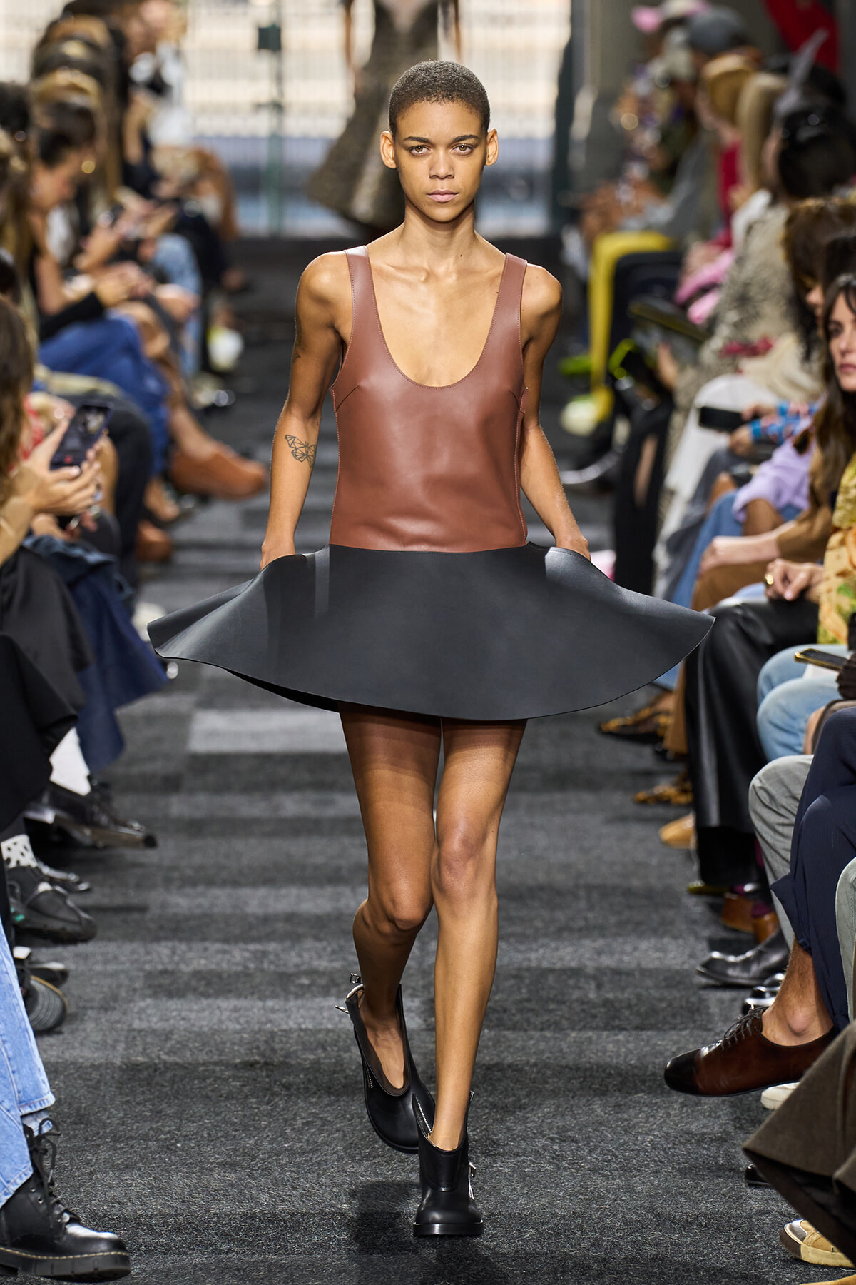 Model walks a fashion runway wearing a brown leather sleeveless top and a flared black skirt, audience on both sides.