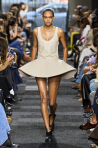 Model walking the runway in a beige sculptural dress with a wide, circular skirt; minimalist sleeveless design and black ankle boots.