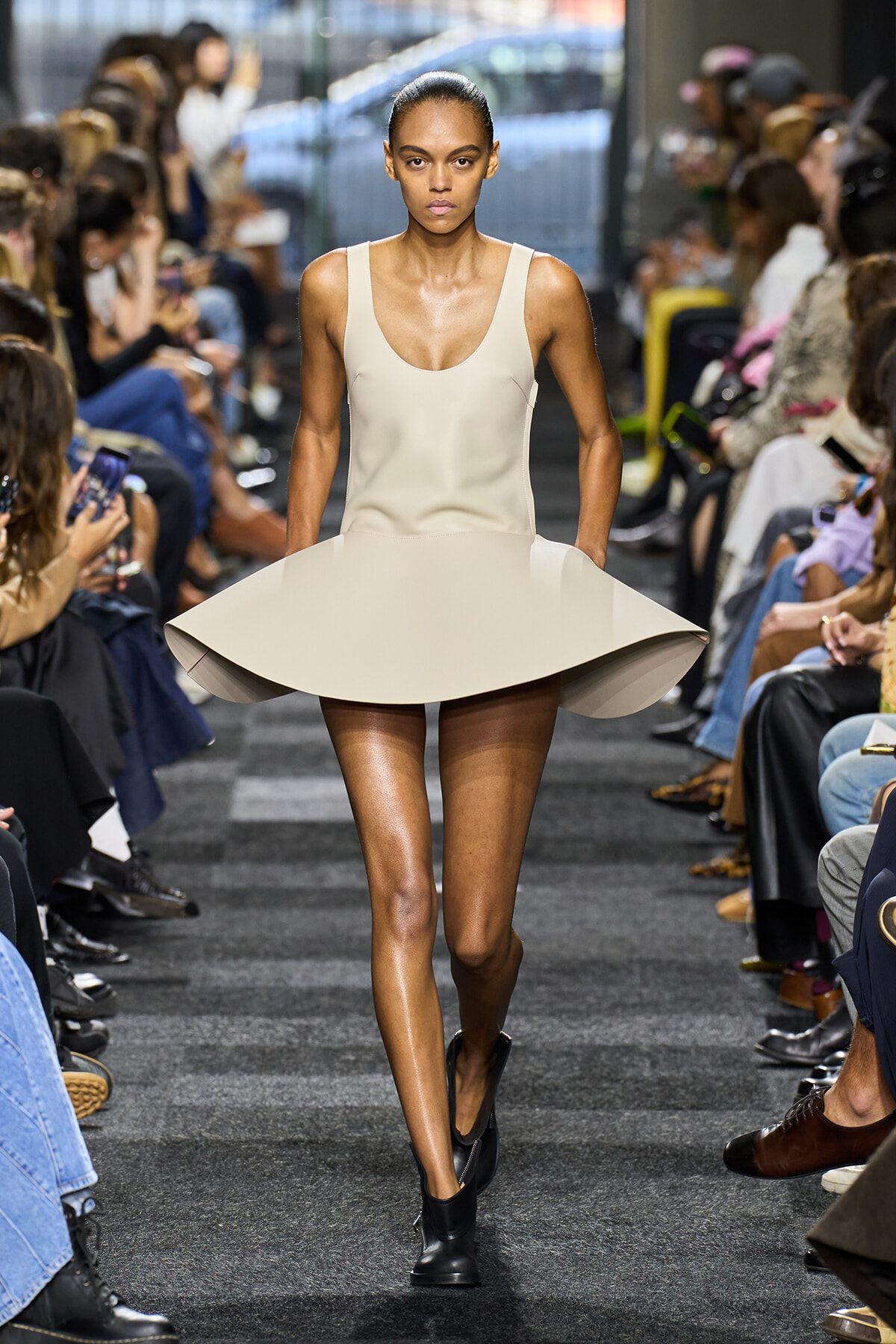 Model walking the runway in a beige sculptural dress with a wide, circular skirt; minimalist sleeveless design and black ankle boots.