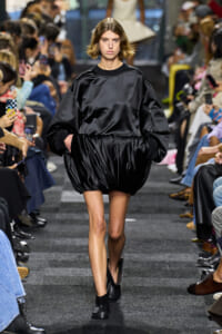 Model walks the runway in an oversized black satin top with a voluminous draped skirt, audience on both sides filming.