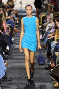 Model walks a bright blue sleeveless knit mini dress down a fashion-show runway, carrying a dark blue handbag.