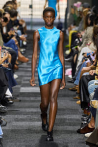 Model walking down the runway in a bright blue sleeveless mini dress, audience on both sides of the catwalk.