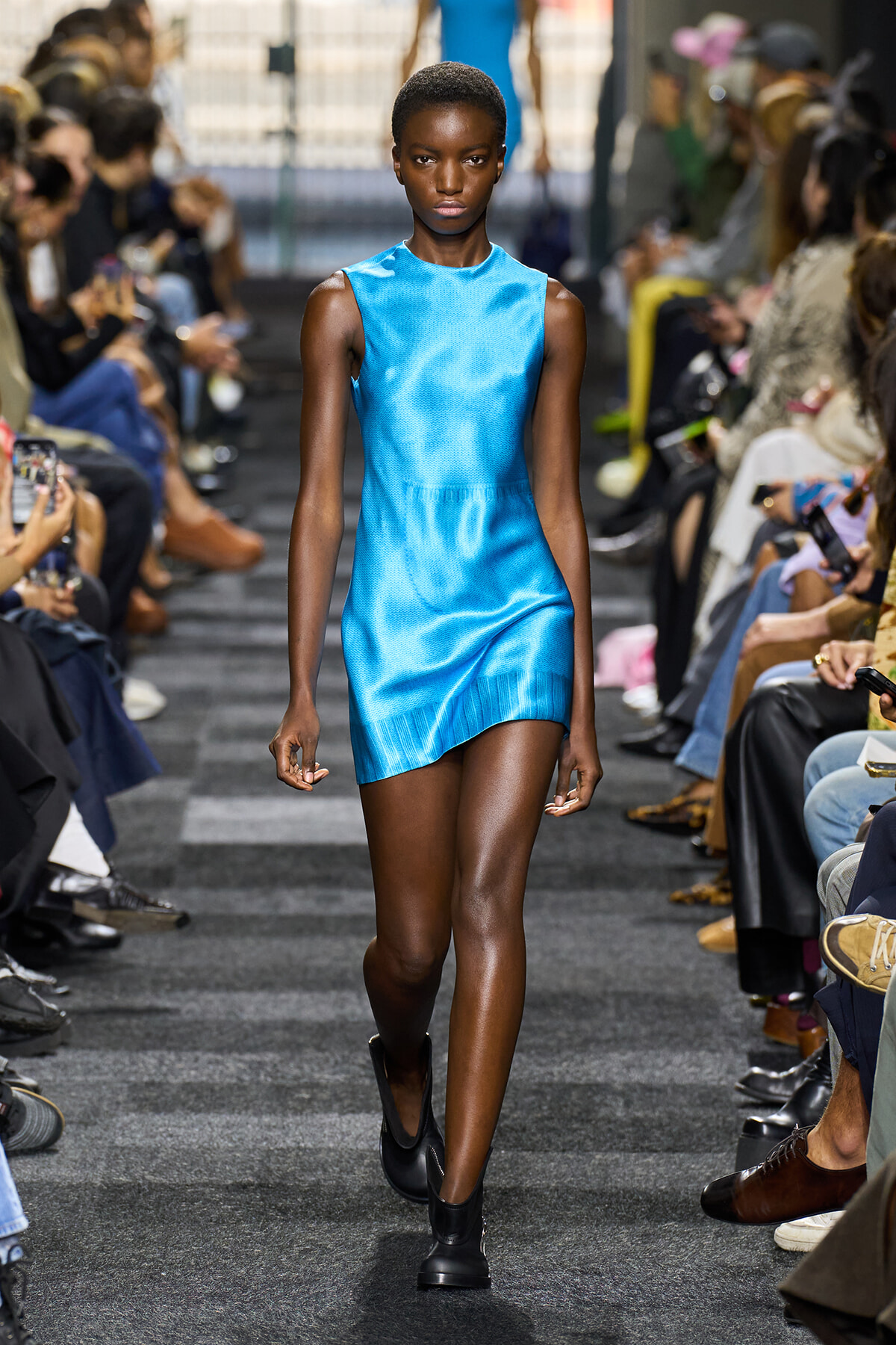 Model walking down the runway in a bright blue sleeveless mini dress, audience on both sides of the catwalk.