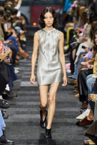 Model walking a fashion runway in a metallic silver sleeveless zip-front mini dress with a hood and drawstrings, paired with black ankle boots; audience watches on both sides of the catwalk.