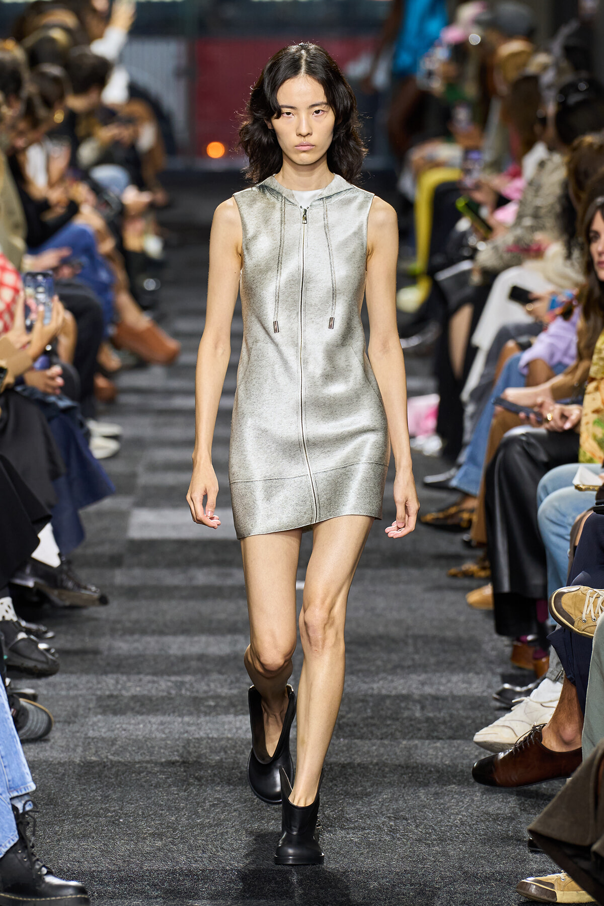 Model walking a fashion runway in a metallic silver sleeveless zip-front mini dress with a hood and drawstrings, paired with black ankle boots; audience watches on both sides of the catwalk.