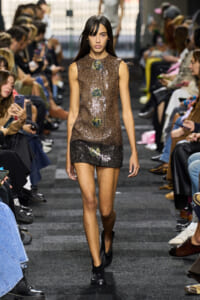 Model walking the runway in a bronze sequin sleeveless dress with circular embellishments, crowds watching on both sides.