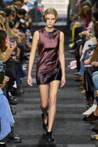 Model walks the runway in a metallic burgundy sleeveless mini dress with oversized black buttons down the front, audience on either side.