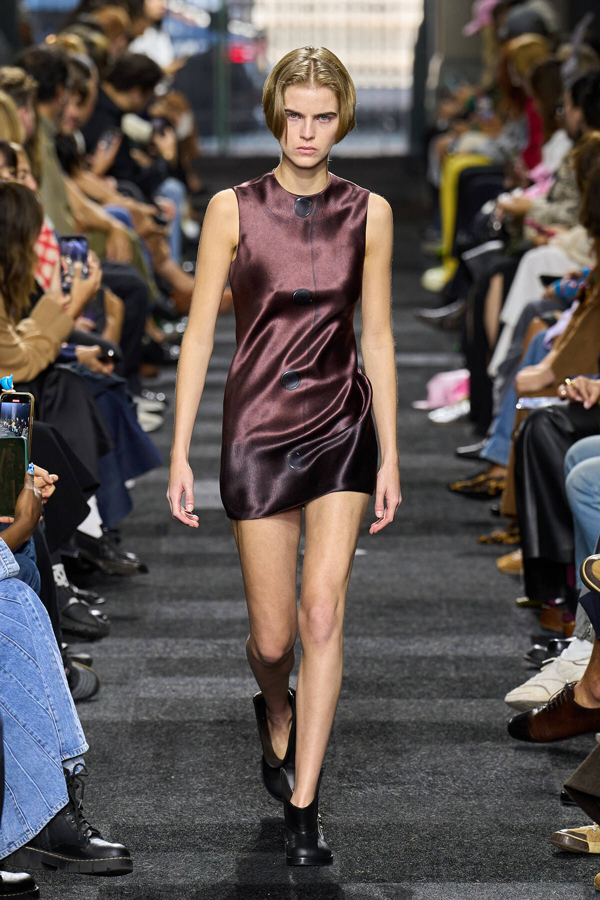 Model walks the runway in a metallic burgundy sleeveless mini dress with oversized black buttons down the front, audience on either side.