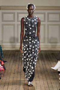 Model on a runway wearing a sleeveless black gown covered in white floral appliqués with a matching floral headpiece on her hair.