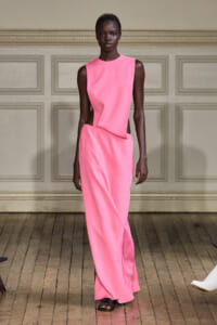Model walking the runway in a bright pink sleeveless jumpsuit with waist cutouts and a draped, asymmetrical waist detail, on a wooden floor.