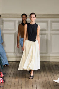 Model walks the runway in a sleeveless black top paired with a high‑waisted, cream pleated midi skirt and black sandals.