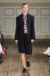 Model walking a fashion runway in an oversized black blazer, pink floral blouse, black knee-length shorts and black slide sandals.