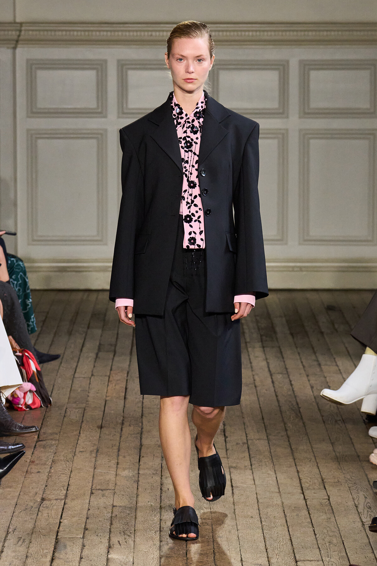 Model walking a fashion runway in an oversized black blazer, pink floral blouse, black knee-length shorts and black slide sandals.