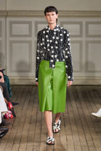 Model on a runway wearing a black floral blouse with white flowers and a bright green midi skirt, paired with floral-embellished sandals.