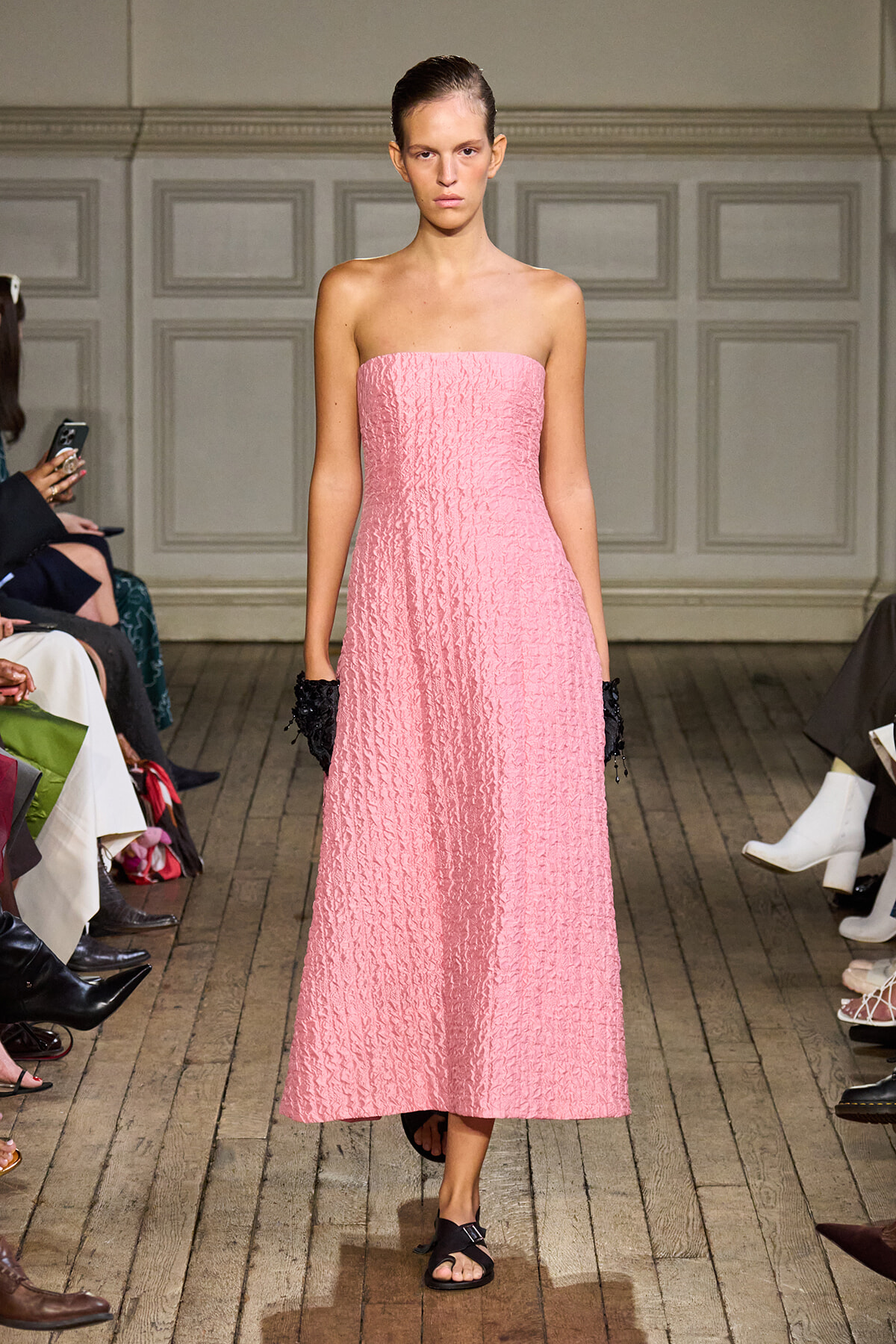 Model walking a runway in a strapless pink textured midi dress, black gloves, and black sandals.