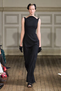 Model walks the runway in a sleeveless black jumpsuit with an asymmetric neckline and black floral headband, gloves, and sandals on a wooden catwalk.