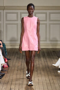 Model walks the runway in a pale pink sleeveless shift dress with a wooden floor and seated audience in the background.