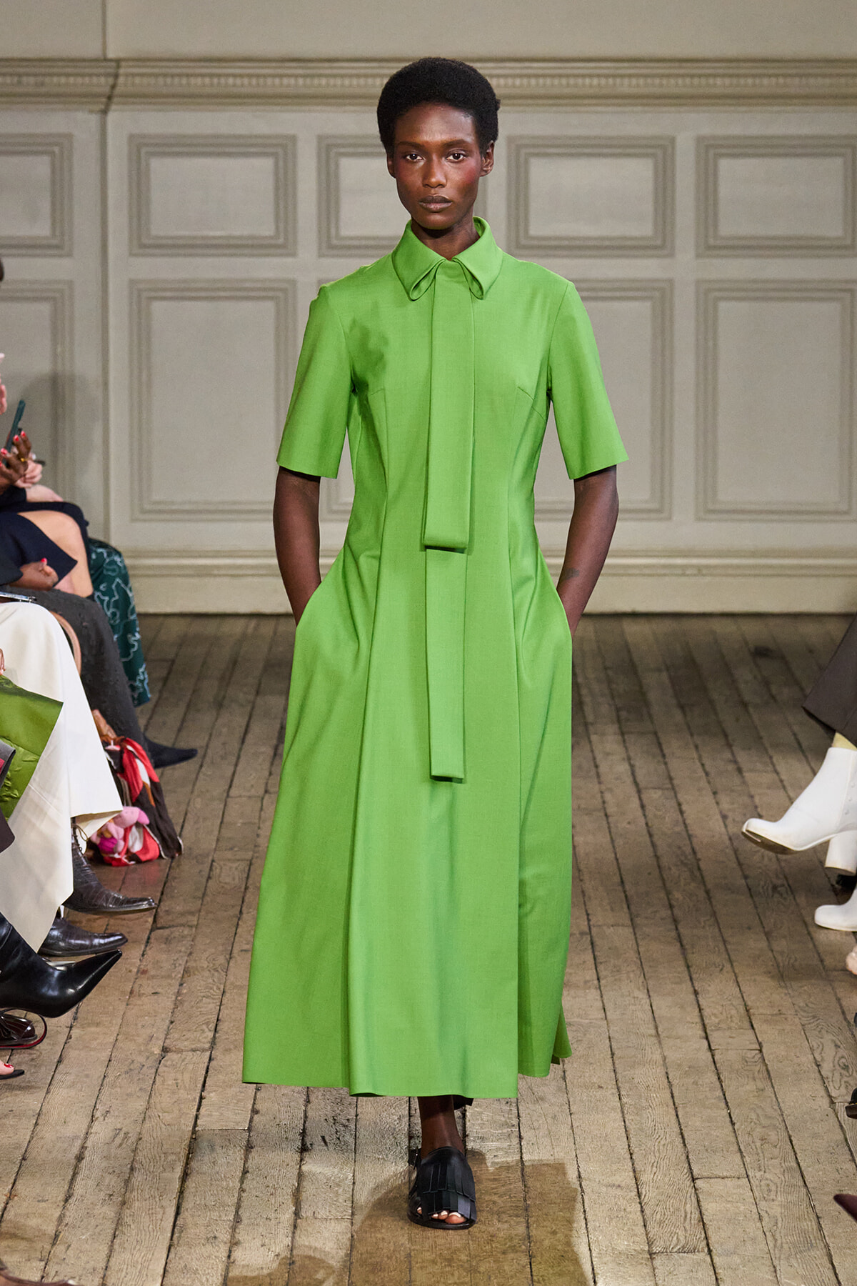 Model on a runway wearing a bright green midi dress with a matching necktie and short sleeves, hands in pockets.