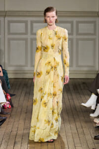 Model walks down a fashion show runway in a pale yellow sheer floral maxi dress with long sleeves.