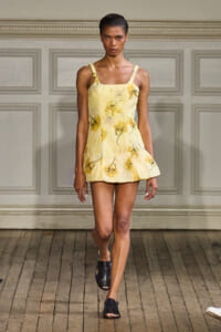 Female model walking a fashion runway in a pale yellow floral sundress with thin straps and black sandals.